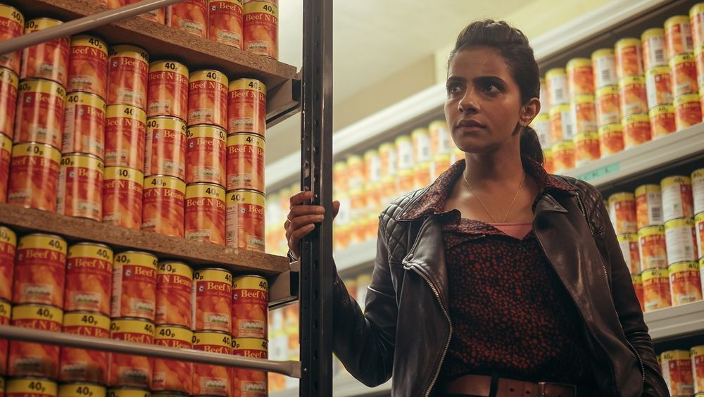 Screenshot of the Doctor Who episode Eve of the Daleks. Yasmin Khan, with a serious look and grabbing one of the metal bar of the front shelf, is standing in a room with lots of shelves, all of them full with 40p cans of beef and beans.