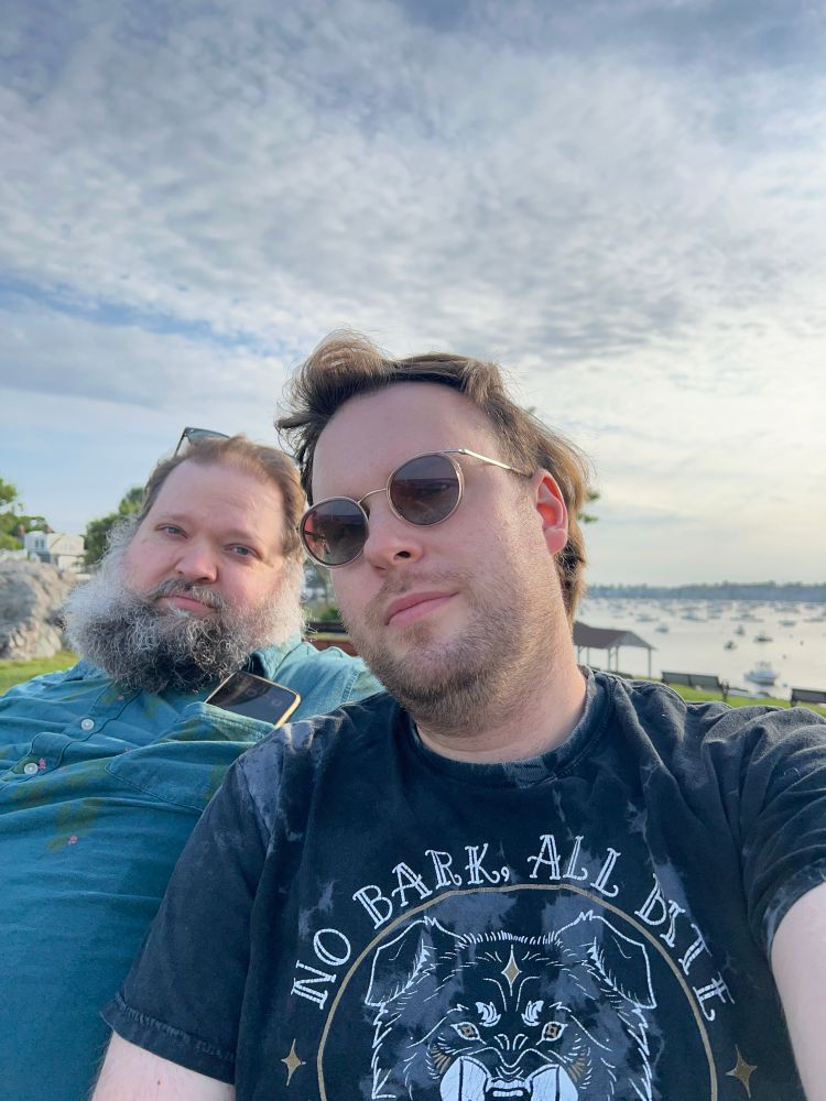Two masc presenting white people, one in the background with a large beard and wearing a blue button up shirt. The other, in the foreground, wearing sunglasses and in a t shirt that says “no bark all bite”
