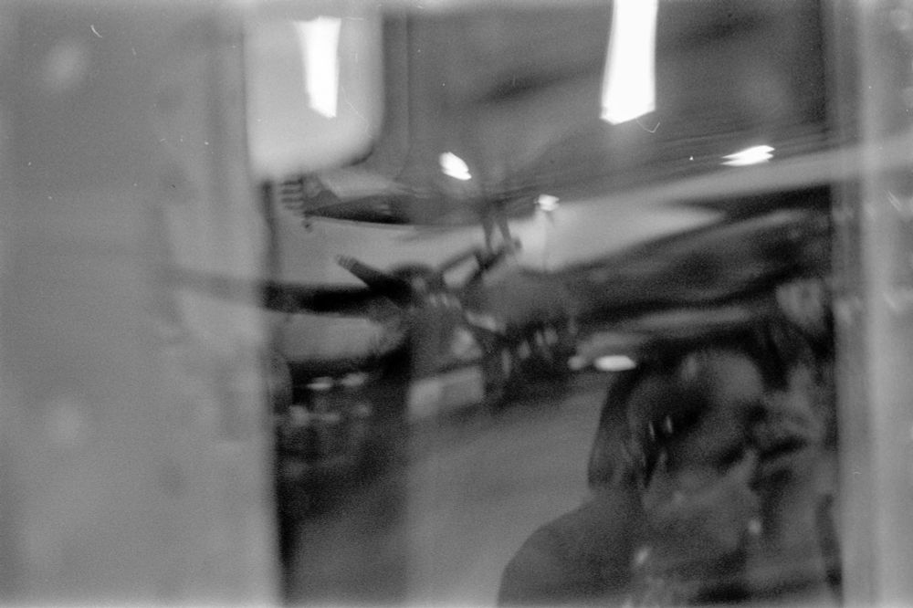 A black & white film image of me reflected in the window of a WWII bomber plane on display in a museum hall. On can also make out the wing and prop of another plane in the background, along with the tail section of a 1940s era fighter plane. I am recognized by the distorted bald patch just above where the camera appears to be, near the bottom right edge of the image.