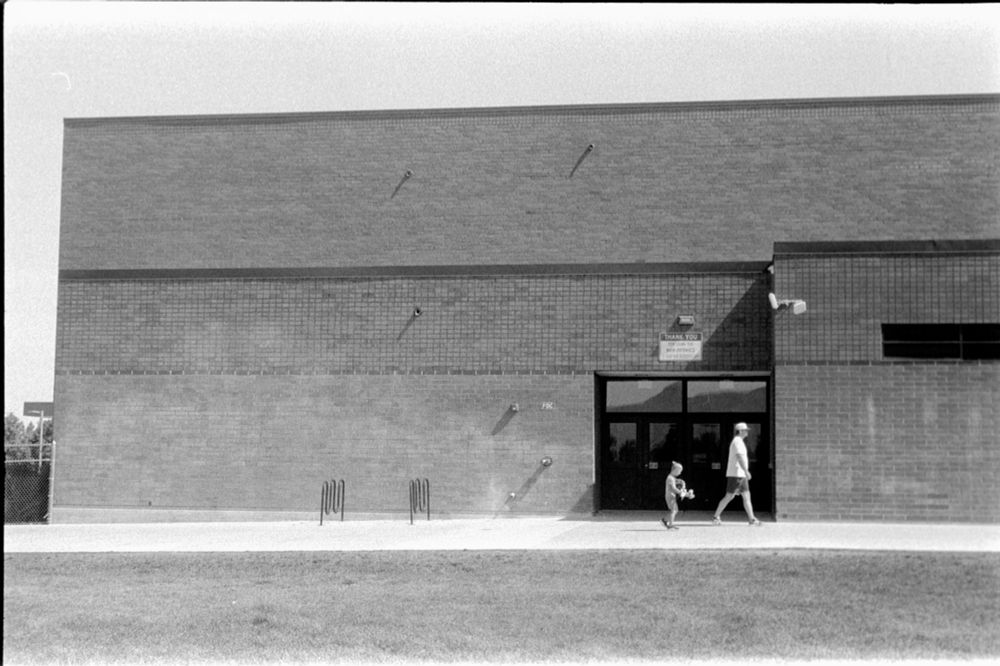 A man and child saunter past a high school building’s door in this black and white photo. 
