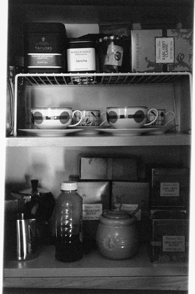 Black and white photo of the inside of a cupboard filled with tea, tea cups, and a honey pot.