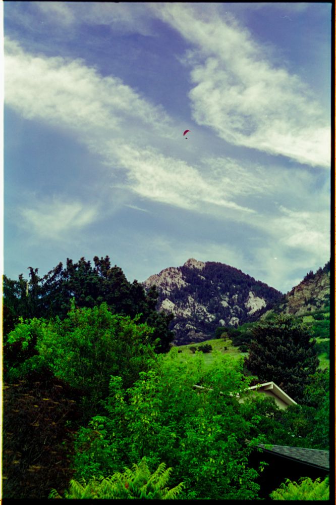 Color photo of trees and a mountain peak in the lower half of the image, with a lightly cloudy sky and a hang glider in the upper half. 