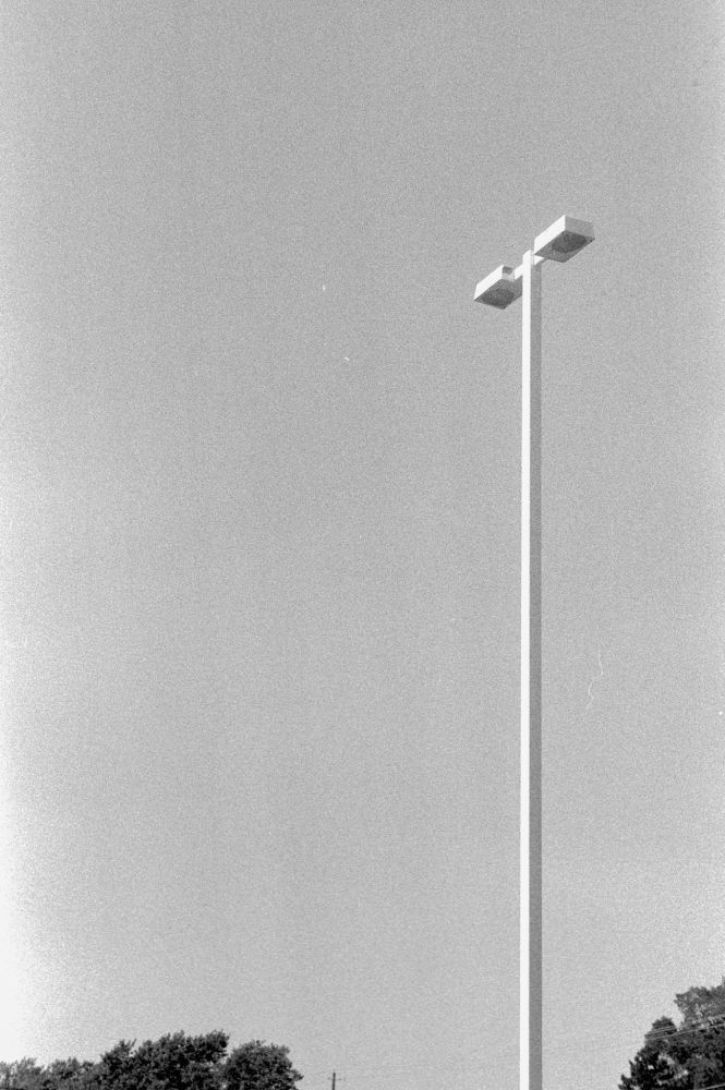 Black & white photo of a parking lot lamp post with just the tops of some trees in view at the bottom of the image.