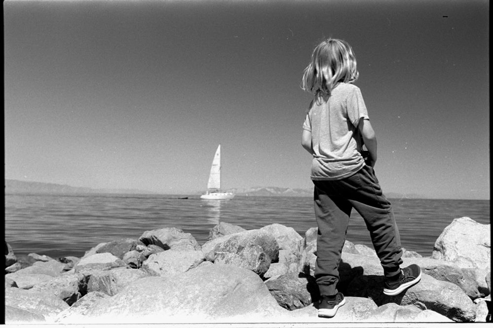 Black and white photo of a boy standing on rocks on the shore, looking out over the lake at a sailboat sailing past. In the distance are mountains that ring the lake. 