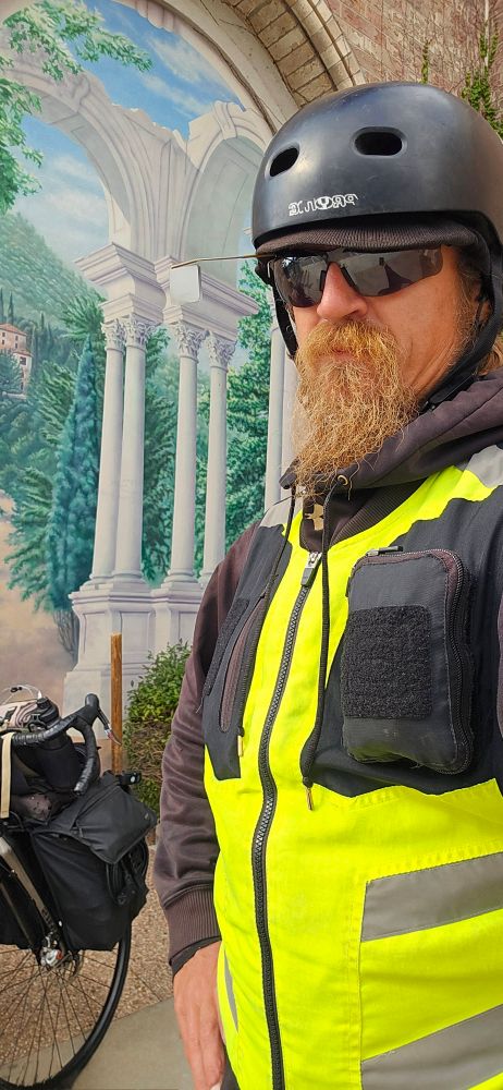 This is a selfie photo of me standing with an artwork depicting roman pillars behind me, and the artwork is within a real archway. My bike is also in the picture in the background.