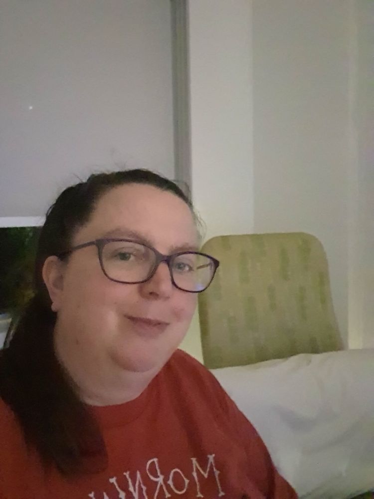 White woman, with glasses and dark hair in a pony tail over one shoulder. She is wearong a red shirt and smiling at the camera. A green chair and white pillow is in the background.