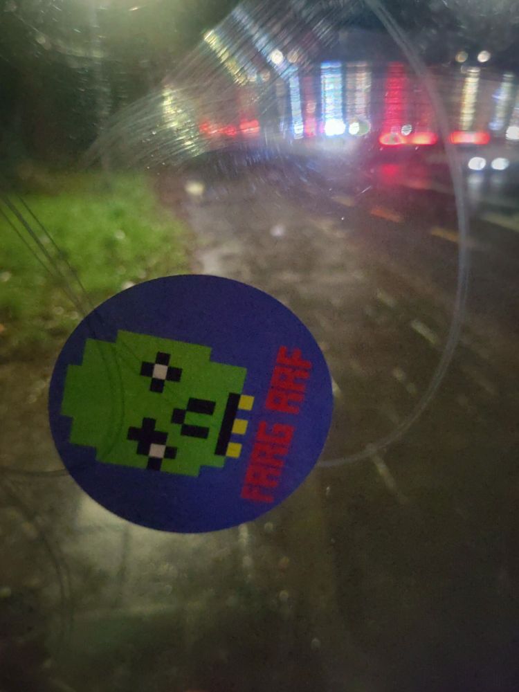 A sticker of a zombie pixelated as it's from Teletext is on a bus shelter window. It's dark and raining and by a busy road (the A406)