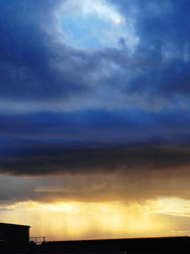 Sky above a black roof with dark blue rain clouds and sun setting a shower of rain is making the yellow horison stripy.