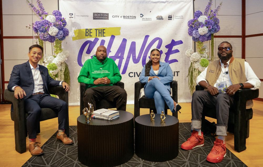 Jerren Chang, Dart Adams, Ebony Gill, and Kenny Mascary sit on stage at the Civic Summit in front of a “Be the Change” banner, flanked by floral arrangements. They are seated in armchairs with small tables between them, holding books and gold decorative figures.