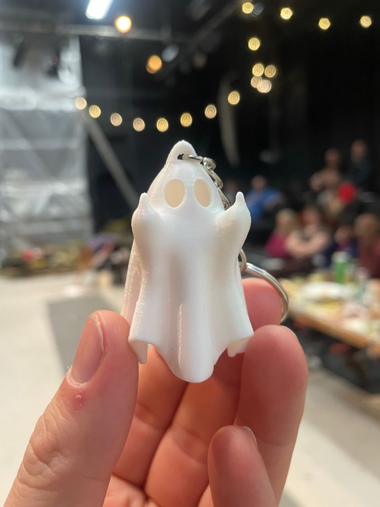A cute little 3D printed ghost throwing up double middle fingers. End of run party out of focus in the background. 