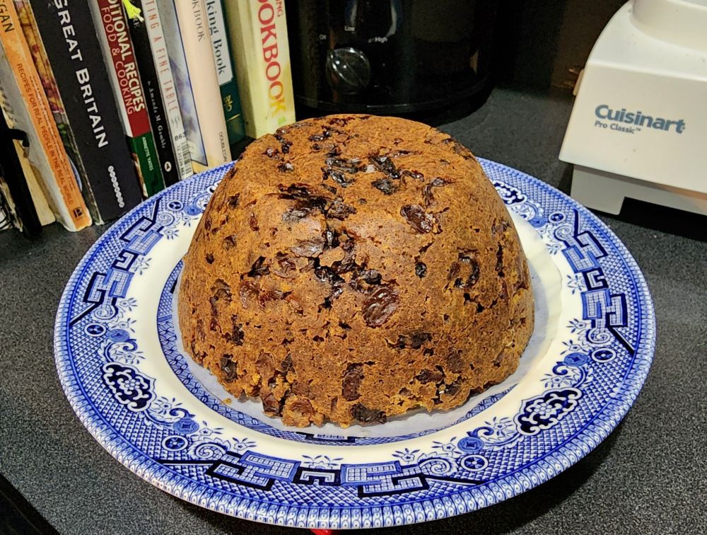 A photograph of traditional Christmas Pudding on a blue willow pattern plate, on my (former) kitchen counter with a backdrop of cookbooks and small appliances. 