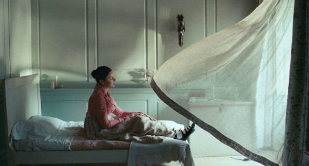 Abbie Cornish sits on a bed, gazing out the window as the sheer curtain billows towards her