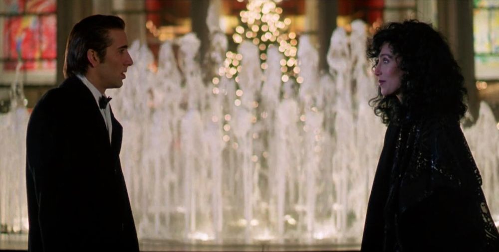 Nicolas Cage & Cher, dressed to the nines, gaze at each other across the frame, in front of the Lincoln Center fountain