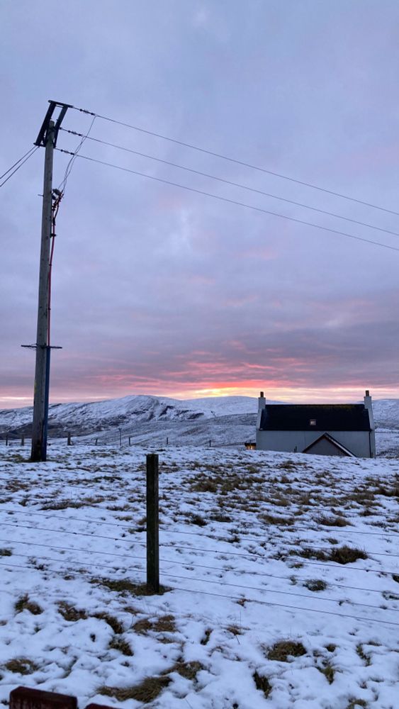 Snowy scene with traditional Shetland house - whitewashed with black tile roof and chimneys at both ends. In the background a bright pink sunset is happening over high snowy hills. 
