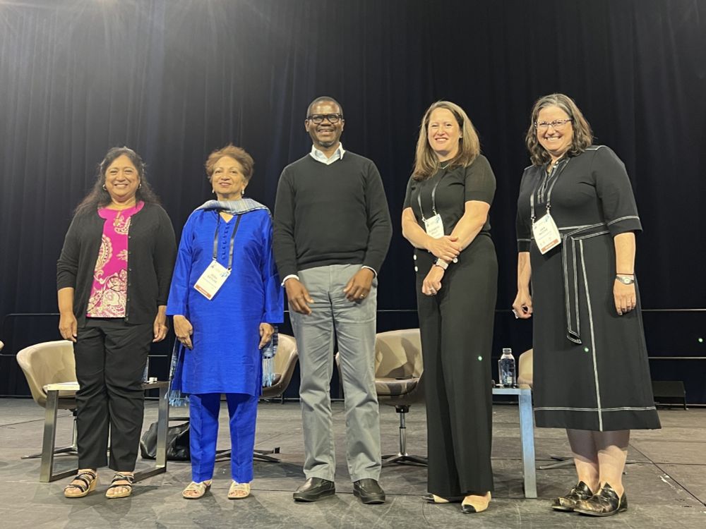 Five speakers are smiling on a stage, with chairs in the background.