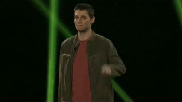 a man in a brown jacket and red shirt is pointing up while standing in front of a green light .