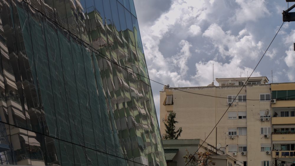Old communism style living blocks are reflected in a modern glass facade in Tirana on a cloudy but sunny day.