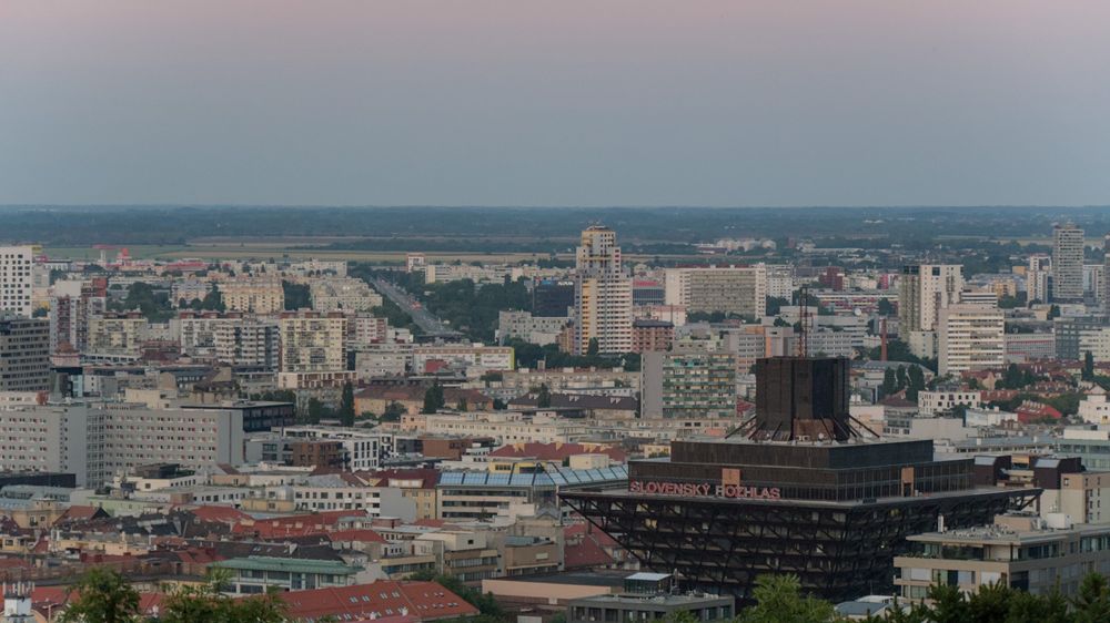 The building of the Slovak state radio as an upturned pyramid on the bottom right of the picture, with high rises and other buildings around it and a straight road angled slightly left leading into the background.