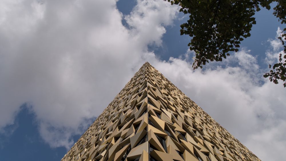 Vor blauen Himmel mit weißen Wolken ragt ein goldenes, viereckiges Gebäude empor. Die Fassade besteht aus goldenen Dreiecken. Am oberen rechten Rand ist auch ein Baumwipfel zu sehen.