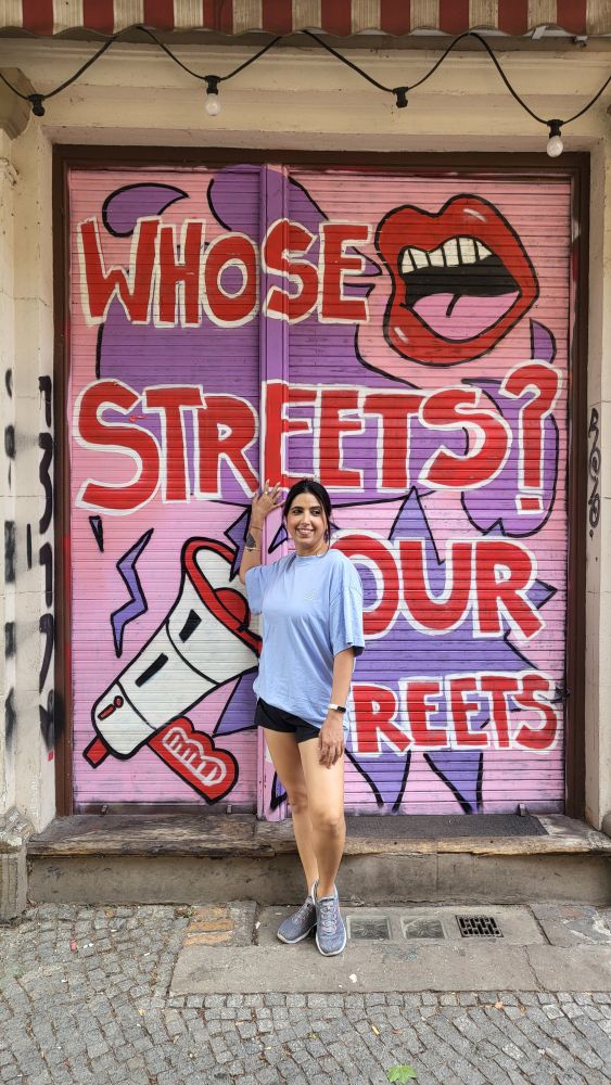 A street art mural featuring text "WHOSE STREETS? OUR STREETS" in red letters on a pink and purple background with a megaphone design.
