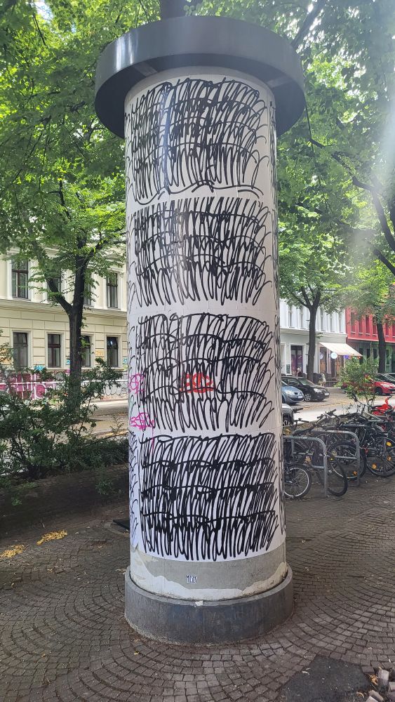 A cylindrical advertising column covered in repetitive black scribble patterns on a white background, located on a street with trees and parked bicycles.