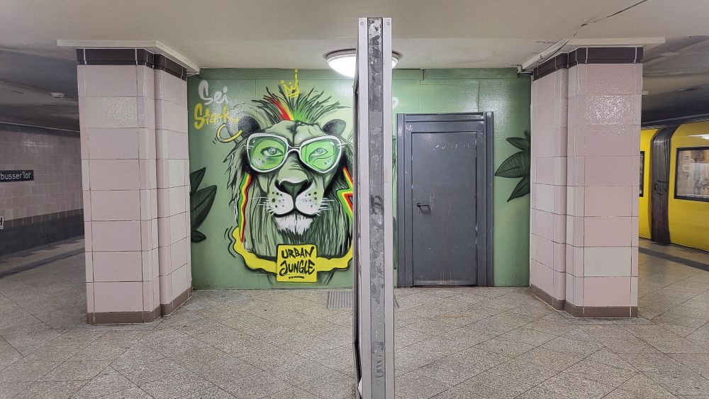 A graffiti mural of a lion wearing green glasses against a mint green background. The lion has a stylized mane and the text "Urban Jungle" beneath it.