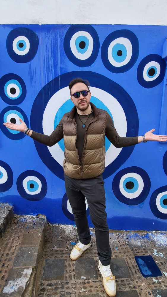 Person standing in front of a blue wall decorated with traditional Turkish 'Nazar' or 'evil eye' symbols in concentric circles of dark blue, white, and light blue. The person is wearing a golden-brown puffer vest, a dark long-sleeve shirt, sunglasses, and is making a welcoming gesture with outstretched arms. He are standing on a cobblestone pavement and wearing cream-colored sneakers.