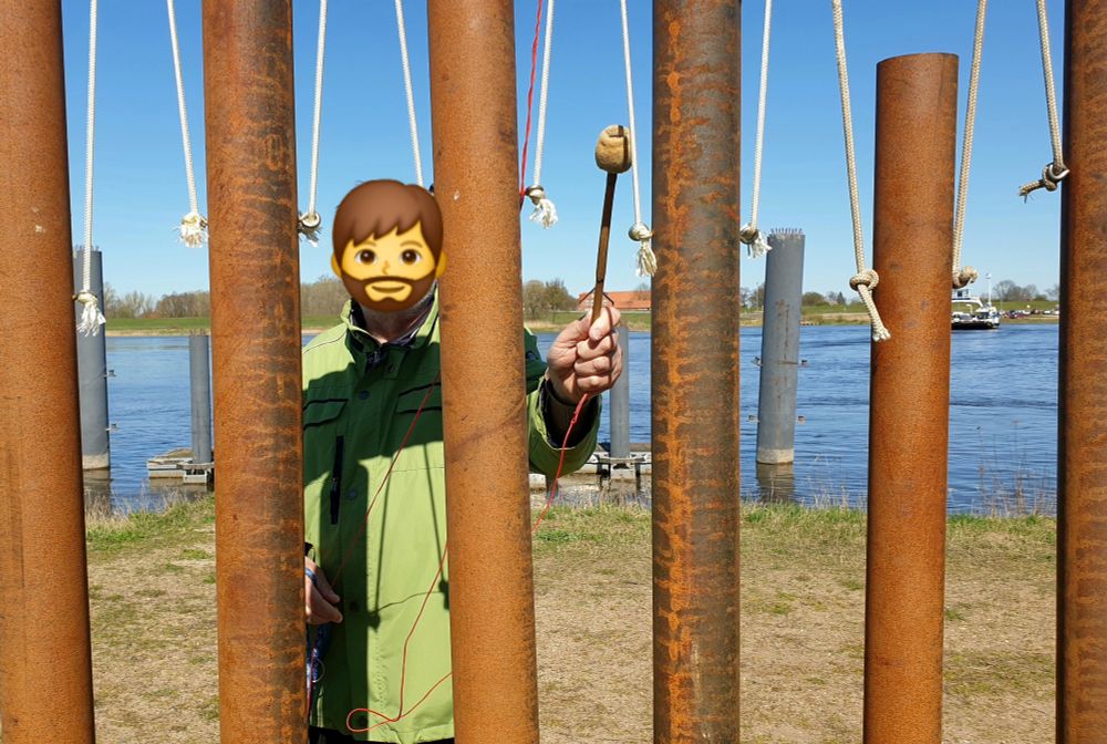Ein großes Klangspiel in der Natur. Man sieht nur die hängenden sechs Röhren, miteinander verbunden mit weißem Seil. Dahinter ein spielenden Mann mit einem Klöppel in der Hand. Im Hintergrund ein Fluss.