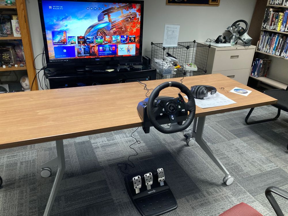 Logitech G920 racing wheel with pedals mounted on a woodgrain table; an Xbox X Home Screen is on a television in the background