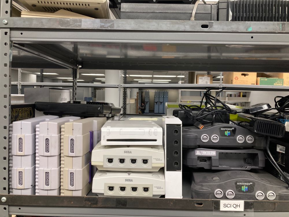 The image shows a shelf filled with vintage video game consoles. On the left, there are several gray Super Nintendo Entertainment System (SNES) consoles stacked together. In the middle, there are two white Sega Dreamcast consoles stacked on top of each other, with a white Nintendo Wii standing next to them and another on top of them. To the right, there are three black Nintendo 64 consoles, with cables resting on and around them. The background shows a large room with shelves and boxes, indicating a storage or collection area.