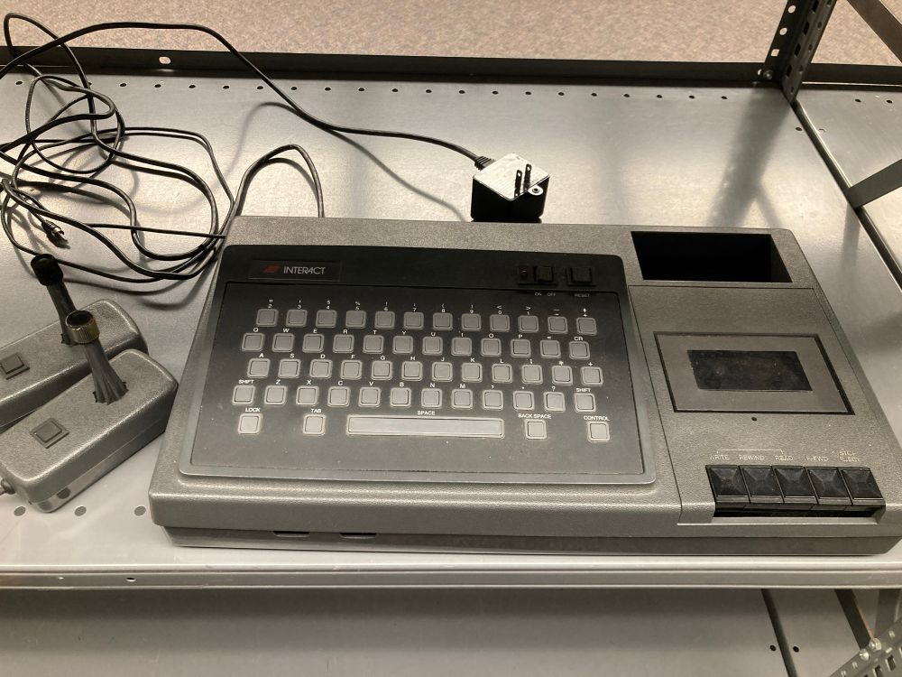 A dark grey Interact Home Computer; with x chiclet keyboard and built-in cassette deck. and two detachable joystick controllers 