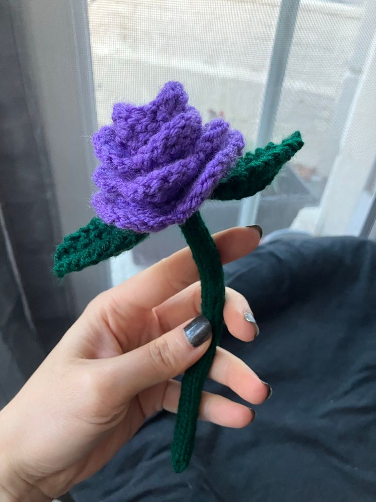 A knitted purple rose with a dark green stem and two leaves. 