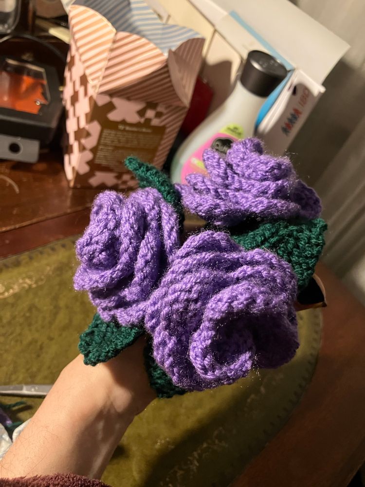 A bouquet of knitted purple roses with a dark green stem and two leaves. 