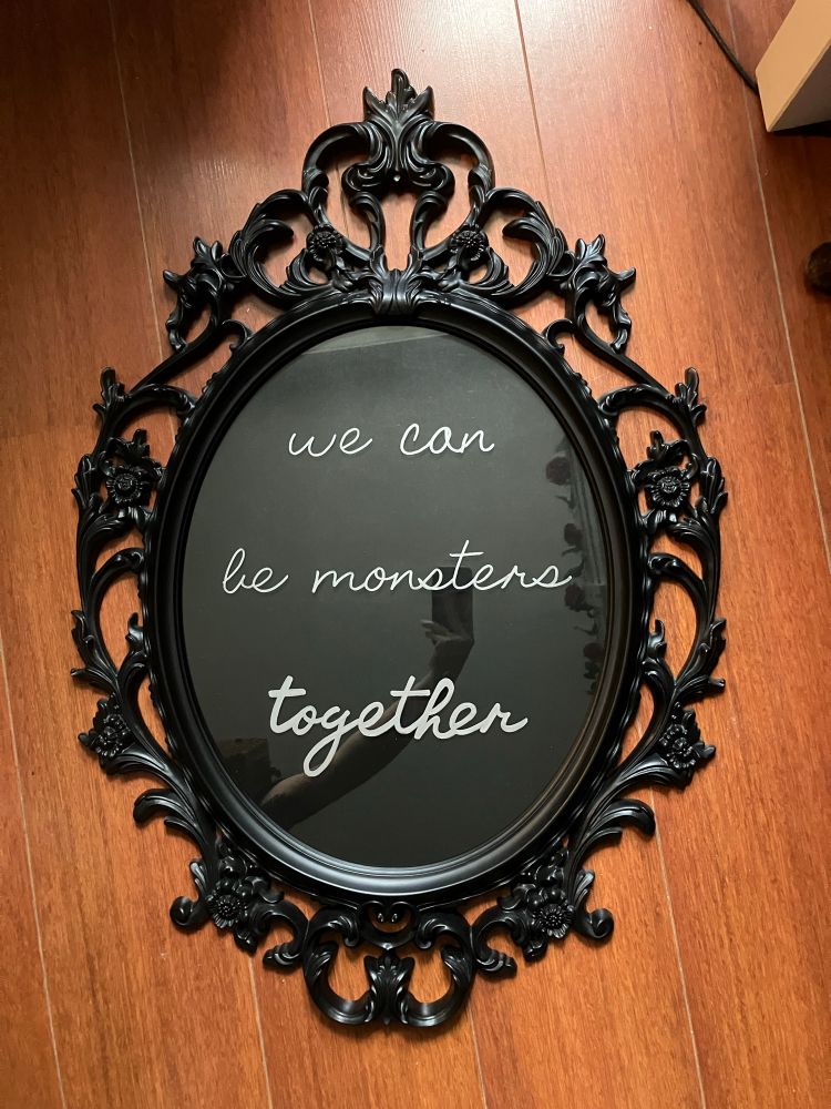 A very ornate black frame (UNG DRILL from IKEA) with a black background inside. The words “we can be monsters together” from the Frankenstein (2025) movie are written on it in cursive with silver paint pen. 