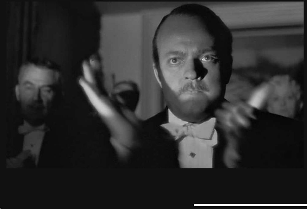 A still from a famous scene in the movie 'Citizen Kane" wherein the mogul, who has been trying to glorify and excuse himself by pushing his wife onto the public, tries to make the audience clap more by standing and clapping wildly