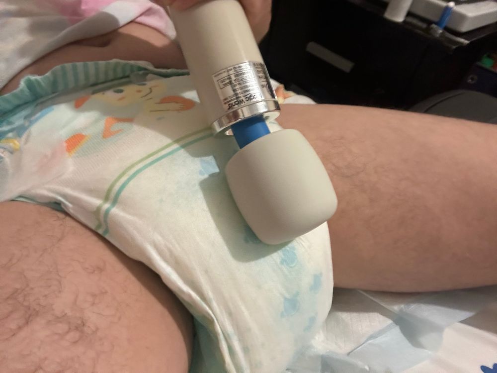 Photo of me wearing a very soggy TryAgains diaper, laying on a puppy pad placed on my bed, pushing a buzzy wand against my front~