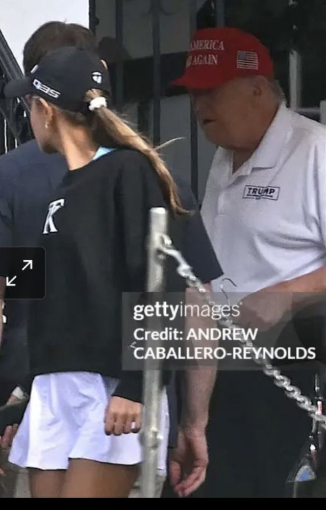 Wider crop of same scene. A woman in front and nearer the camera than turnip has a shaggy blonde ish brown ish ponytail thing on her shoulder. The depth of field in the photo is distorted, the ponytail looks larger and closer to him than it actually is. 