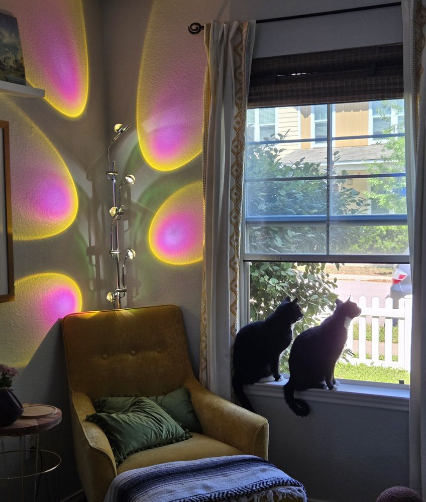 Picture of the corner of a living room, a yellow chair and a colorful lamp  sits behind it. Two Felix cats sit on the windowsill next to the chair