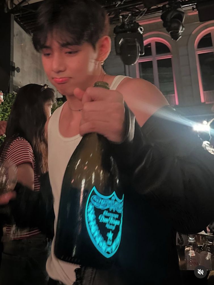 taehyung holding a bottle of champagne in a club or a restuarent in paris. He’s wearing a white tank top with a leather jacket that is hanging off one shoulder