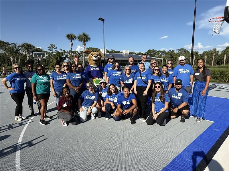 A group of people wearing "KPMG" shirts posing with a kangaroo mascot on a basketball court under clear skies.