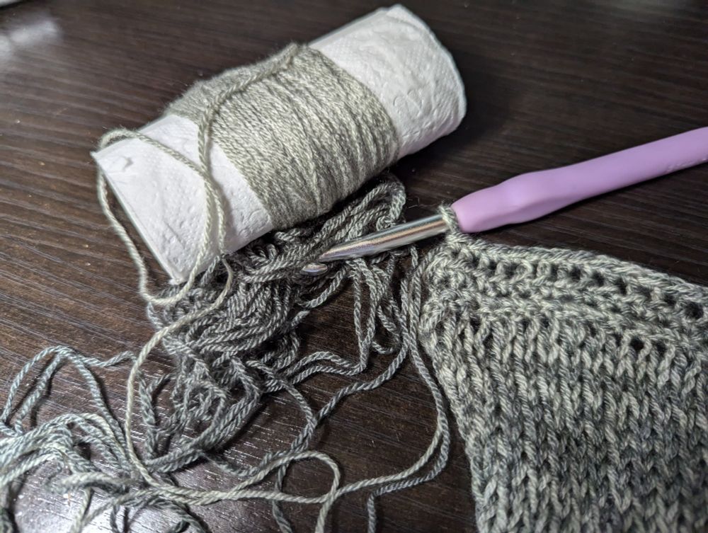 End of a row of crochet border on a knit panel. Remaining unused yarn on a toilet paper roll and in a small pile. 