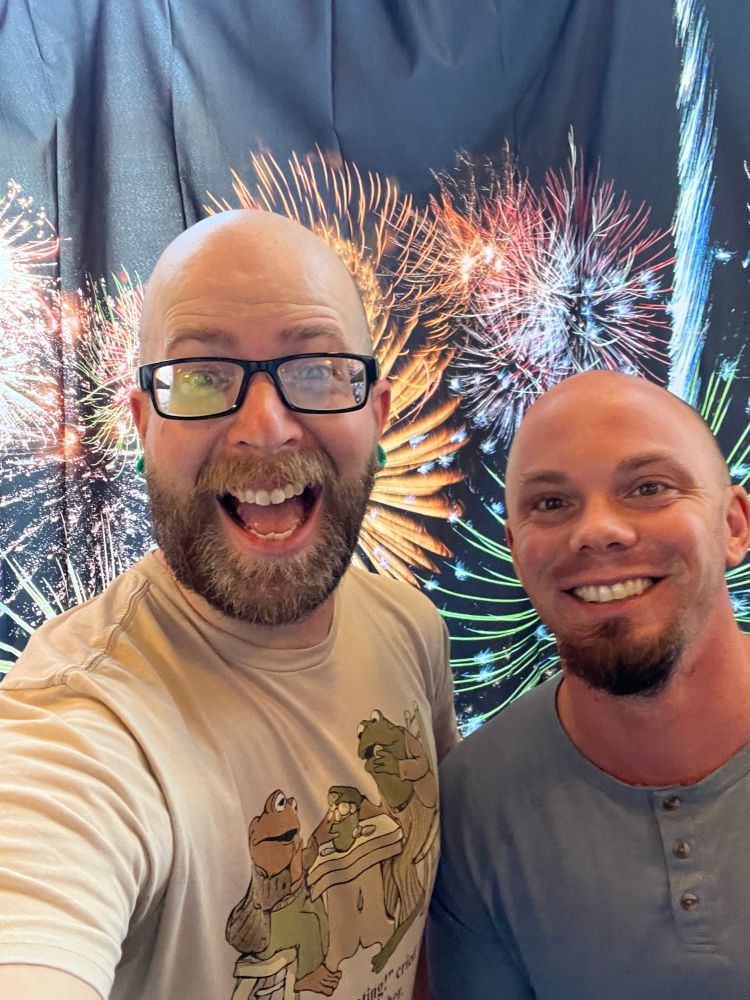 Two bald gentlemen excited to be on front of a fireworks backdrop together.