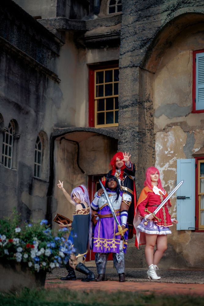 A cosplay photo taken at a castle. There are windows with red frames, flowers in the front, and a few old arches. The Tailtiu cosplayer is in her promoted outfit with a black cape and cold decorations, holding a Thoron tome and pointing left as if casting. Arvis stands in the back holding out his hand. Ayra stands in front with her sword, and Ethlyn stands on the right looking out while holding her sword.