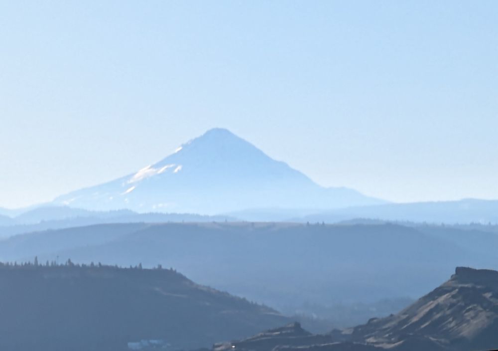 A photo of Mount Hood (also known as Wy'east).  A light blues/gray vista of the mountain in the backdrop with forest and hillscapes closer to the camera.  Blue sky.