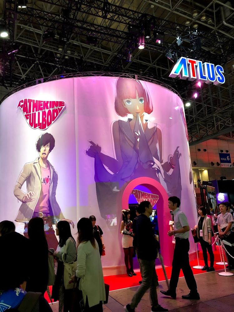 A character in Catherine Full Body holds open a dress, serving as an entrance to a booth at a game conference event