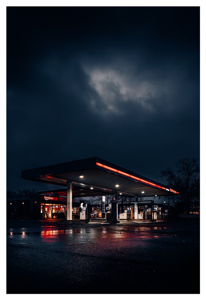 A gas station at night with dark clouds, rainy ground and red lights.