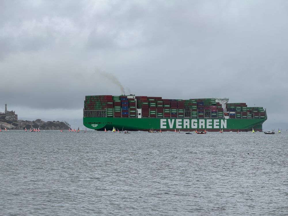 Huge green cargo ship entering San Francisco Bay. Alcatraz in the background. Dozens of small sailboats surround the ship 