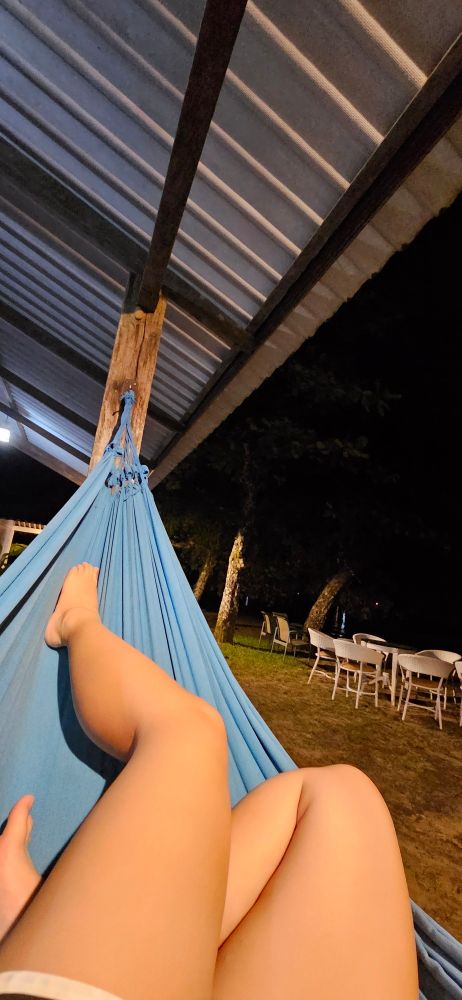 pernas em uma rede azul. ao fundo se vê um gramado com cadeiras e palmeiras 