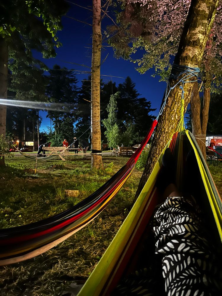 Resting on a hammock with some festivees in the background. It is dark, the area is green and surrounded by trees. 