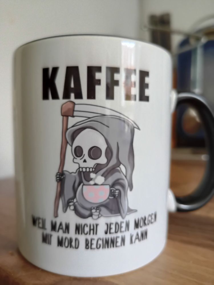 Kaffeebecher. Mittig aufgedruckt eine Zeichnung vom Tod: Skelett in langem Gewand mit Kaputte, hält eine Sense und eine große rosa Tasse mit türkisen Punkten.
Text darüber: Kaffee
Text darunter: Weil man nicht jeden Morgen mit einem Mord beginnen kann